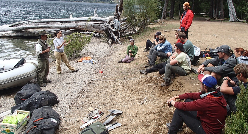 A group of people sit on the shore, listening to two people speak. In the water, there is a raft.